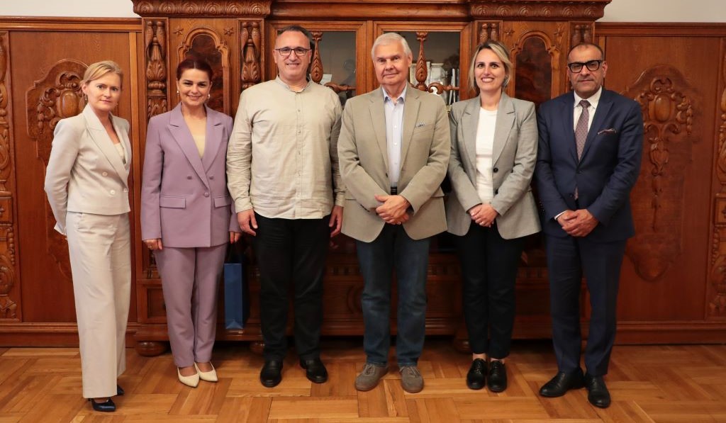 Six people pose for a group photo in a formal interior, against the background of decorative wooden furnishings. The participants stand in one row and look towards the camera.