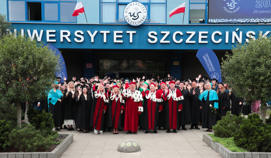 A group of representatives of the University of Szczecin in ceremonial academic gowns stands in front of the university building. In the background, the inscription “University of Szczecin”, the US logo, Polish flags and university banners are visible.